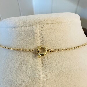 K&ouml;nnte beinhalten: Eine goldfarbene Halskette mit einer filigranen Kette und einem runden Verschluss. Die Halskette wird auf einer wei&szlig;en Schaufensterpuppe pr&auml;sentiert, um ihr Design hervorzuheben. Der Verschluss ist schlicht und elegant.