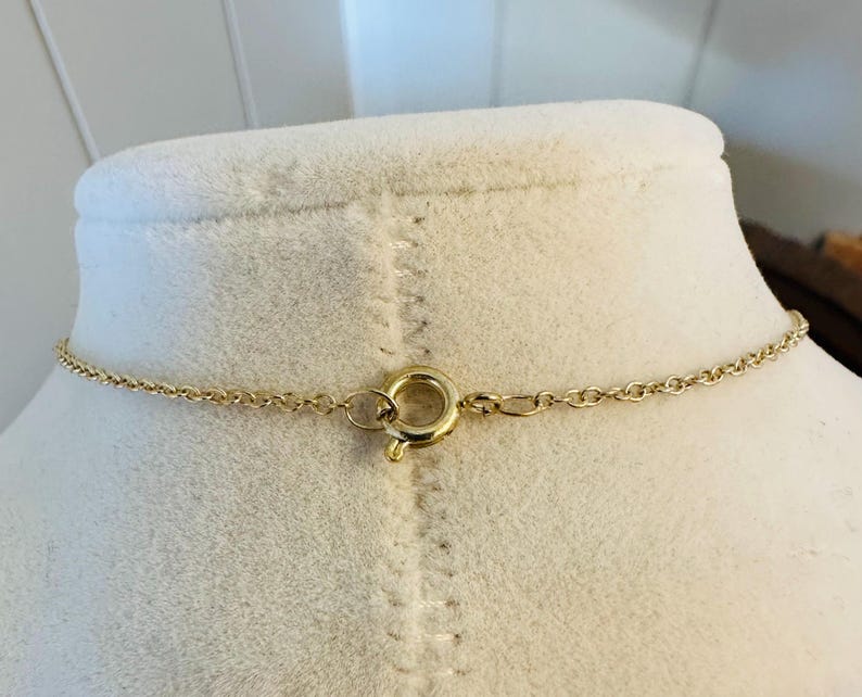 K&ouml;nnte beinhalten: Eine goldfarbene Halskette mit einer filigranen Kette und einem runden Verschluss. Die Halskette wird auf einer wei&szlig;en Schaufensterpuppe pr&auml;sentiert, um ihr Design hervorzuheben. Der Verschluss ist schlicht und elegant.