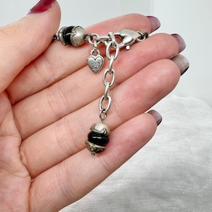 Puede incluir: Un collar de cadena de plata con un dije de coraz&oacute;n y detalles de cuentas negras. La cadena tiene un cierre de langosta.