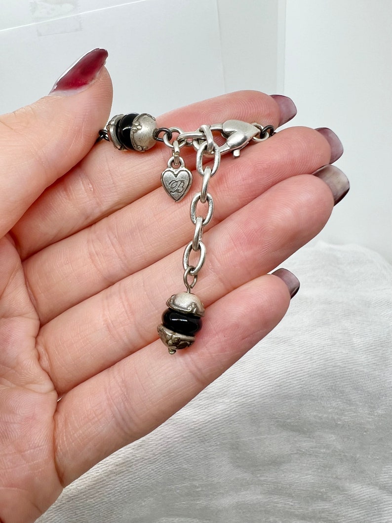 Puede incluir: Un collar de cadena de plata con un dije de coraz&oacute;n y detalles de cuentas negras. La cadena tiene un cierre de langosta.