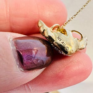 K&ouml;nnte beinhalten: Eine goldfarbene Anh&auml;ngerhalskette mit einem stilisierten Design. Der Anh&auml;nger hat eine strukturierte Oberfl&auml;che und ist mit kleinen, funkelnden Akzenten verziert. Die Kette ist zart und erg&auml;nzt das Design des Anh&auml;ngers. Der Anh&auml;nger ist mit dem Text ".925 AU" markiert.