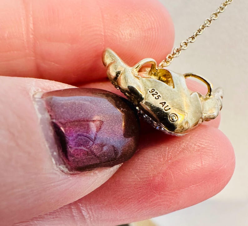 K&ouml;nnte beinhalten: Eine goldfarbene Anh&auml;ngerhalskette mit einem stilisierten Design. Der Anh&auml;nger hat eine strukturierte Oberfl&auml;che und ist mit kleinen, funkelnden Akzenten verziert. Die Kette ist zart und erg&auml;nzt das Design des Anh&auml;ngers. Der Anh&auml;nger ist mit dem Text ".925 AU" markiert.