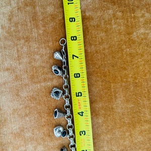 May include: Silver charm bracelet with various charms, including a skull, a heart, and a ring. The bracelet is approximately 7 inches long, as measured by a yellow ruler.