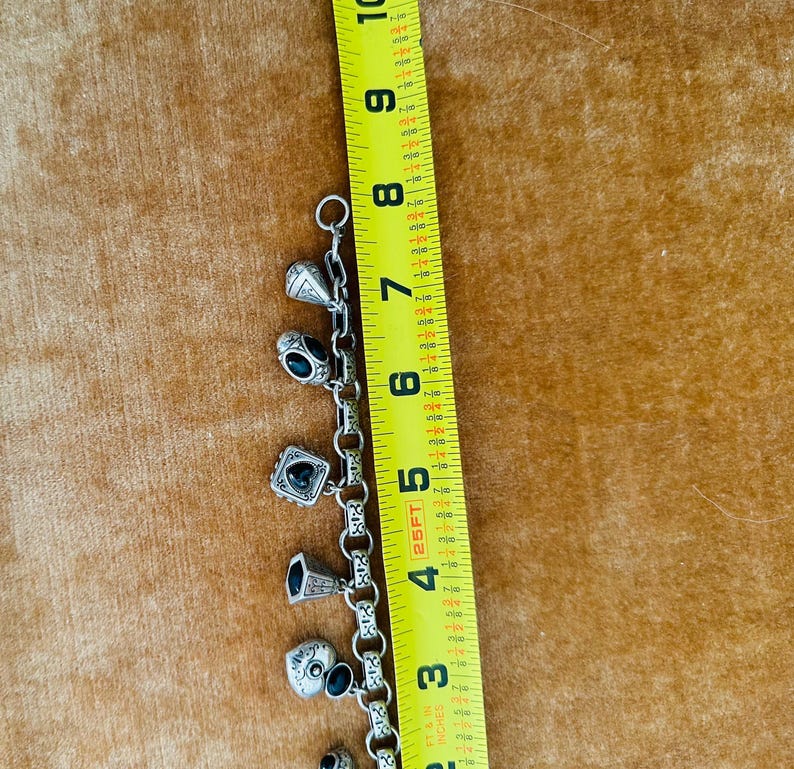 May include: Silver charm bracelet with various charms, including a skull, a heart, and a ring. The bracelet is approximately 7 inches long, as measured by a yellow ruler.