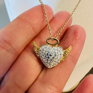 K&ouml;nnte beinhalten: Goldfarbene Halskette mit herzf&ouml;rmigem Anh&auml;nger. Der Anh&auml;nger hat einen Heiligenschein und Fl&uuml;gel. Das Herz ist mit wei&szlig;en Kristallen besetzt, Fl&uuml;gel und Heiligenschein sind goldfarben.