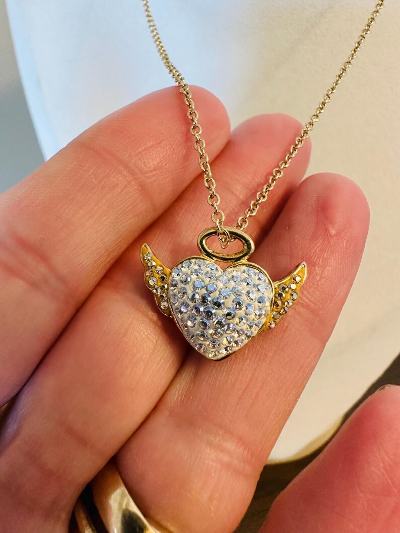 K&ouml;nnte beinhalten: Goldfarbene Halskette mit herzf&ouml;rmigem Anh&auml;nger. Der Anh&auml;nger hat einen Heiligenschein und Fl&uuml;gel. Das Herz ist mit wei&szlig;en Kristallen besetzt, Fl&uuml;gel und Heiligenschein sind goldfarben.