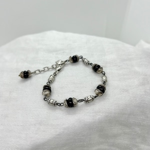 Puede incluir: Un brazalete de plata con cuentas negras y un cierre de plata. El brazalete est&aacute; sobre un fondo de tela blanca.