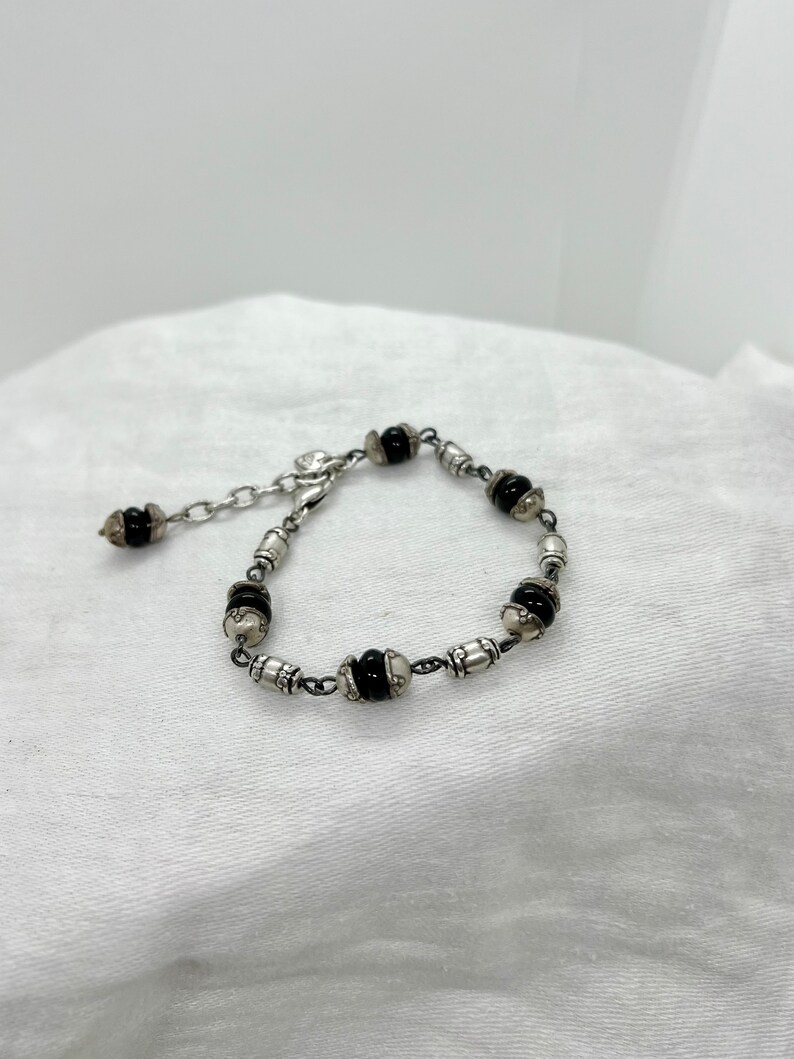 Puede incluir: Un brazalete de plata con cuentas negras y un cierre de plata. El brazalete est&aacute; sobre un fondo de tela blanca.