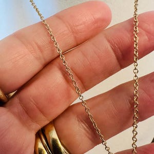 K&ouml;nnte beinhalten: Nahaufnahme einer zarten Goldkettenhalskette. Die Kette besteht aus kleinen, miteinander verbundenen Gliedern, die ein feines und elegantes Aussehen erzeugen. Die warme Goldfarbe kontrastiert mit dem Hintergrund und hebt das filigrane Design des Schmucks hervor. Die Halskette wird in einer Hand gehalten.