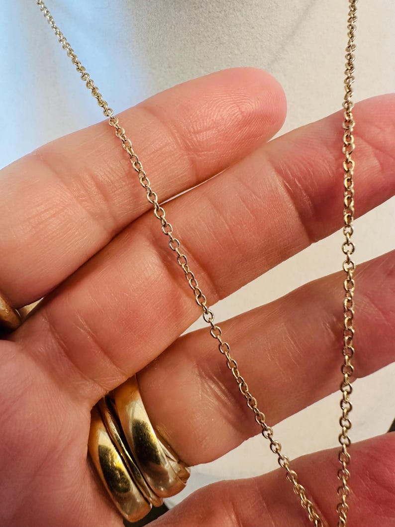 K&ouml;nnte beinhalten: Nahaufnahme einer zarten Goldkettenhalskette. Die Kette besteht aus kleinen, miteinander verbundenen Gliedern, die ein feines und elegantes Aussehen erzeugen. Die warme Goldfarbe kontrastiert mit dem Hintergrund und hebt das filigrane Design des Schmucks hervor. Die Halskette wird in einer Hand gehalten.