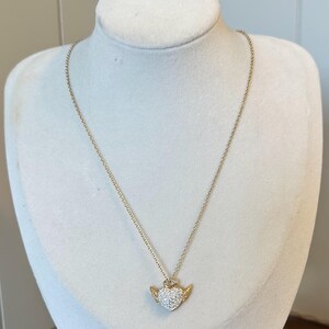 K&ouml;nnte beinhalten: Eine goldfarbene Halskette mit einem herzf&ouml;rmigen Anh&auml;nger. Das Herz ist mit kleinen, klaren Kristallen besetzt und hat kleine, goldfarbene Fl&uuml;gel. Die Halskette wird auf einer wei&szlig;en Schaufensterpuppe pr&auml;sentiert.