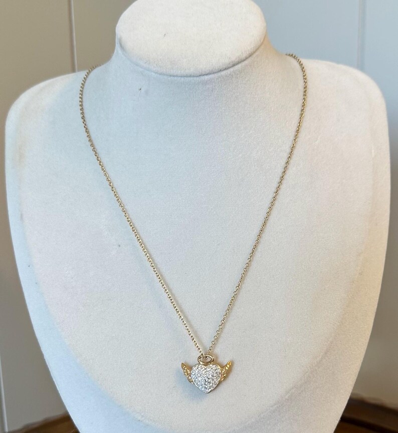 K&ouml;nnte beinhalten: Eine goldfarbene Halskette mit einem herzf&ouml;rmigen Anh&auml;nger. Das Herz ist mit kleinen, klaren Kristallen besetzt und hat kleine, goldfarbene Fl&uuml;gel. Die Halskette wird auf einer wei&szlig;en Schaufensterpuppe pr&auml;sentiert.