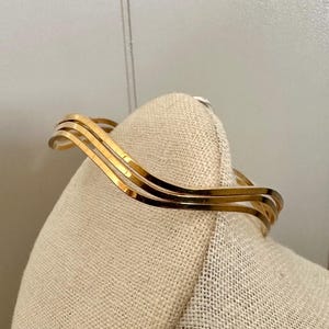 May include: A gold-toned bracelet with a wavy design. The bracelet features four parallel, curved bands that create a flowing, elegant look. The jewelry is displayed on a beige fabric surface.