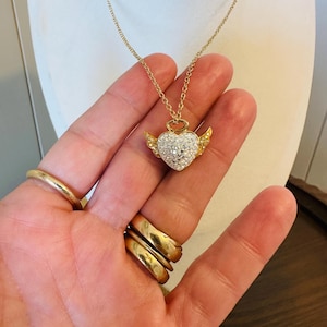 K&ouml;nnte beinhalten: Eine goldfarbene Halskette mit einem herzf&ouml;rmigen Anh&auml;nger. Das Herz ist mit kleinen, klaren Kristallen besetzt und hat kleine goldfarbene Fl&uuml;gel. Die Halskette wird auf einer wei&szlig;en Mannequin-B&uuml;ste pr&auml;sentiert.