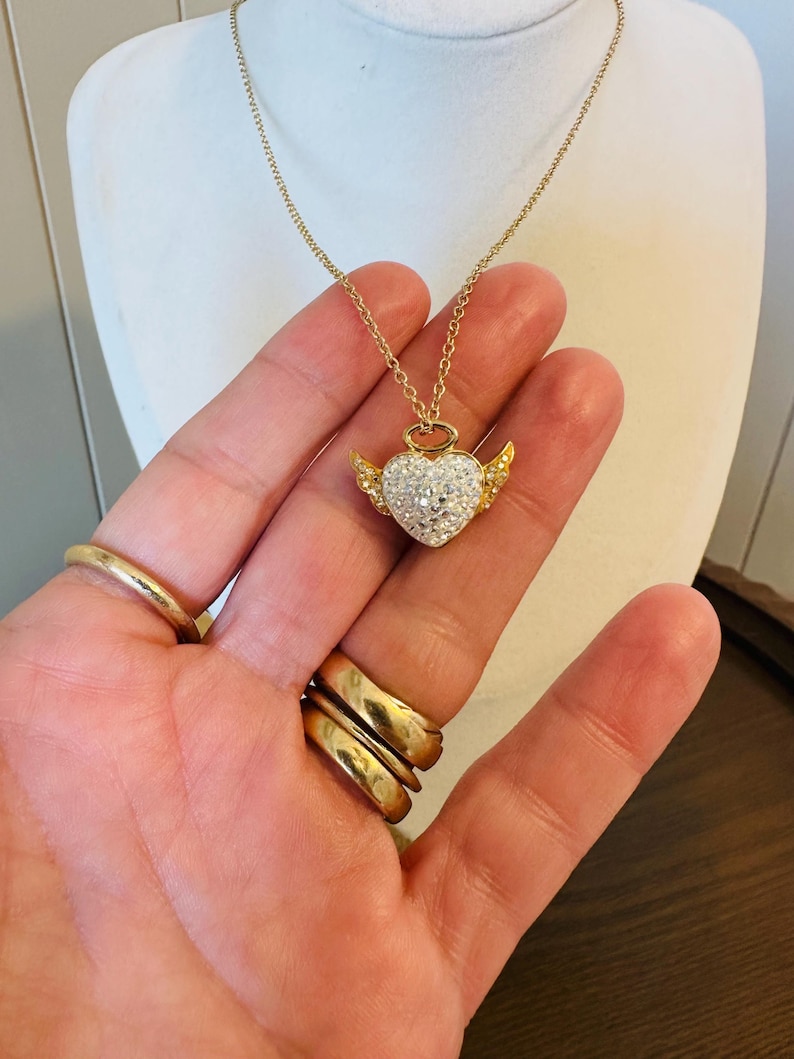 K&ouml;nnte beinhalten: Eine goldfarbene Halskette mit einem herzf&ouml;rmigen Anh&auml;nger. Das Herz ist mit kleinen, klaren Kristallen besetzt und hat kleine goldfarbene Fl&uuml;gel. Die Halskette wird auf einer wei&szlig;en Mannequin-B&uuml;ste pr&auml;sentiert.