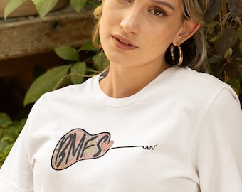 May include: A woman wearing a white t-shirt with a guitar graphic and the text "BMFS" on it. She is also wearing a white skirt, a brown belt with a large metal buckle, and several gold bracelets. She is sitting on a set of stairs with a potted plant behind her.
