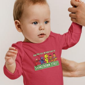 May include: A red baby bodysuit with long sleeves. The front displays a graphic of Sesame Street characters and the text "CAN YOU TELL ME HOW TO GET TO SHAKEDOWN STREET". The baby is being held by an adult.