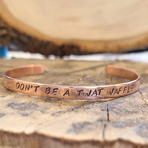 May include: A copper bracelet with the inscription "Don't be a twat waffle" on a wooden surface.