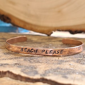 Puede incluir: Una pulsera de cobre con las palabras "Beach Please" grabadas. La pulsera está sobre una superficie de madera.