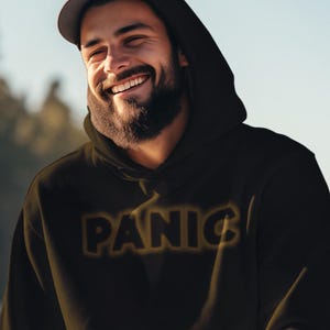 May include: Black hoodie with the word "PANIC" in a gold outline. The hoodie has a drawstring hood and a front pocket. The person is wearing a black cap and tan pants.