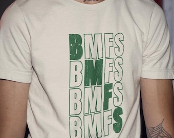 May include: Cream-colored t-shirt with the green text "BMFS" stacked vertically. The shirt is short-sleeved and appears to be made of a soft material. The overall style is casual and modern.