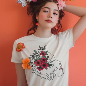 May include: A woman wearing a white t-shirt with a floral design and the text "Not like other girls." She has a flower crown and butterfly accents in her hair. The background is a solid orange.