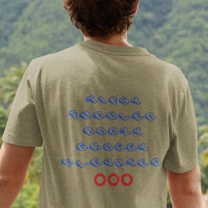 May include: A light olive green t-shirt with blue text that reads "TIPSY PUDDLES BOOZY ELEVATED" and three red circles. The person is standing outside with a mountain view.