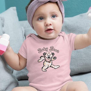 May include: A baby wearing a pink onesie with the words "Baby Bear" and a cartoon bear graphic. The baby is also wearing a pink headband and holding pink and white toy blocks.