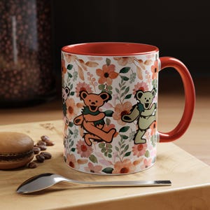May include: A red and white ceramic mug with a floral pattern and three dancing bears. The bears are orange, green, and brown. The mug has a red rim and handle.