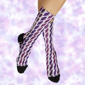 May include: A pair of knee-high socks with a repeating zigzag pattern in shades of purple and white. The socks have black heels and toes. The background is a soft, blurred purple and white.