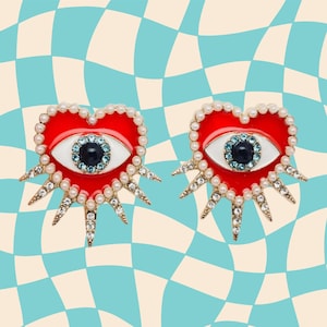 May include: A pair of heart-shaped earrings with a red enamel base, outlined with faux pearls. Each earring features a white eye with a blue gem center and gold-tone starburst accents. The earrings are set against a blue and cream checkered background.