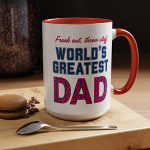 May include: A white ceramic mug with a red interior and handle. The mug features the text "Freak out, throw stuff" above "WORLD'S GREATEST DAD" in blue and red lettering. A spoon and a macaron are on a wooden surface.