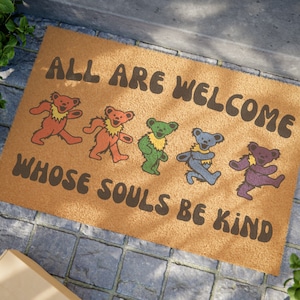 May include: A brown doormat with the text "All are welcome whose souls be kind" in a retro font. Five colorful dancing bears are depicted on the mat.