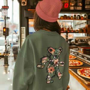 Könnte beinhalten: Olivgrünes Sweatshirt mit einem floralen Bärenmuster auf dem Rücken. Der Bär ist mit einem bunten Blumenmuster gefüllt. Eine rosa Mütze wird getragen. Der Hintergrund zeigt eine Restaurantkulisse mit Pizza.