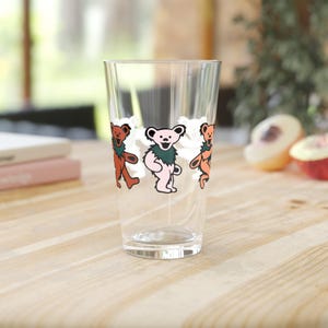 May include: A clear glass pint glass with a design of four dancing bears. The bears are orange, pink, and brown with green accents.