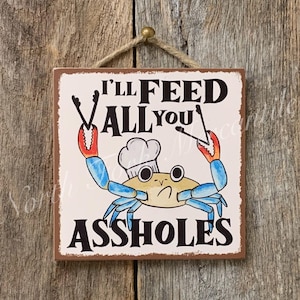 May include: Square wooden sign with a crab chef illustration and the text "I'LL FEED ALL YOU ASSHOLES". The crab is light yellow with blue claws and a chef's hat. The sign is hanging on a wooden background.