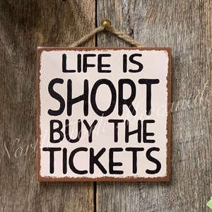 May include: A white wooden sign with brown edges and black text that reads "Life is Short Buy the Tickets". The sign is hanging from a piece of twine with a gold metal hook.