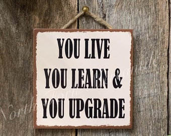 Puede incluir: Un letrero de metal blanco con bordes marrones colgado en una pared de madera. El letrero dice "You live you learn & you upgrade".