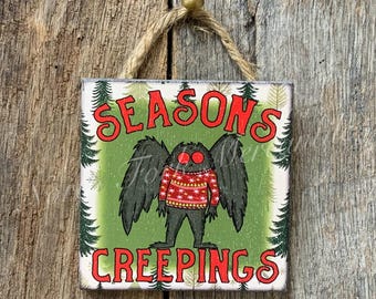 May include: Square wooden sign with the text "SEASONS CREEPINGS" in red. The sign features a cartoon monster with black wings and a red and white sweater. The background is green with white accents and a rope hanger.