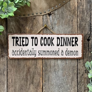 May include: A rustic brown metal sign with white text that reads "Tried to cook dinner accidentally summoned a demon".