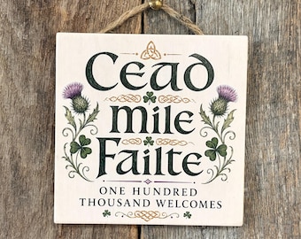 May include: A square wooden sign with a light cream background featuring the dark green text Cead mile failte and the English translation One hundred thousand welcomes. The design includes purple thistle illustrations and green shamrocks.