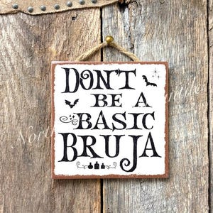 May include: Square wooden sign with the text "Don't Be A Basic Bruja" in black lettering. The sign has a white background with a brown border and is hanging from a jute rope. The background is a rustic wooden surface.