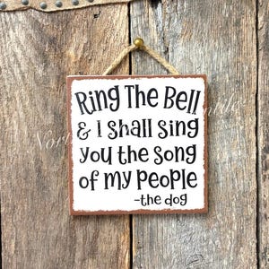 May include: Square wooden sign with the text "Ring The Bell & I shall sing you the song of my people -the dog" in black font. The sign has a brown border and is hanging from a jute rope. The background is a rustic wooden wall.