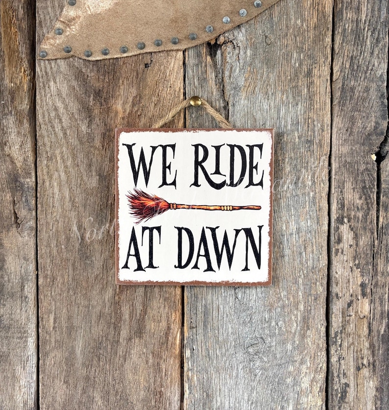 May include: Square wooden sign with the text "WE RIDE AT DAWN" in black, with a broom graphic. The sign is hanging from a jute rope and is set against a rustic wooden background. The sign is a decorative item.