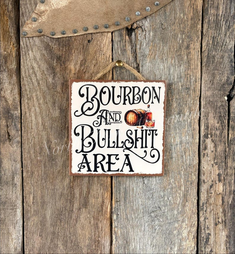 Peut inclure: Panneau carr&eacute; en bois avec les mots "Bourbon and Bullshit Area" en &eacute;criture noire. Le panneau pr&eacute;sente une illustration d'un tonneau de bourbon et d'un verre de bourbon. Le panneau est accroch&eacute; &agrave; un mur en bois rustique.