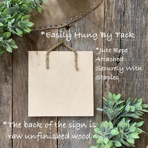 May include: Square wooden sign with jute rope hanger. The sign is light-colored with visible wood grain. Text on the sign reads, "Easily Hung By Tack," "Jute Rope Attached Securely with Staples," and "The back of the sign is raw unfinished wood."