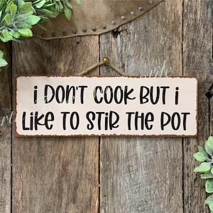 May include: A rustic metal sign with a white background and black text that reads "i don't cook but i like to stir the pot". The sign is hanging from a piece of twine and is surrounded by green plants.