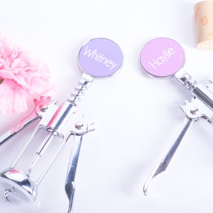 May include: Two silver wine bottle openers with personalized round tops, one lavender and one pink, each with a name printed on it. A pink flower and a cork are also in the image.