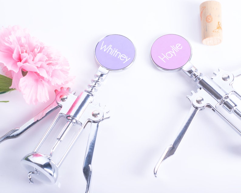 May include: Two silver wine bottle openers with personalized round tops, one lavender and one pink, each with a name printed on it. A pink flower and a cork are also in the image.
