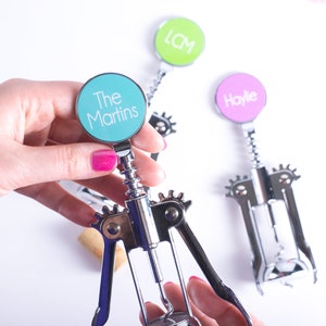 May include: Silver wine bottle opener with a teal circle that says "The Martins". Two other openers are visible, one with "LCM" in green and another with "Haylie" in pink. The openers have a corkscrew design and are held by a person with pink nail polish.